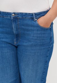 Jeans de ganga azul com um corte ajustado, apresentando um design de cinco bolsos e um pequeno fecho de botão metálico na cintura. Textura suave.