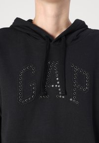 Felpa nera con cappuccio caratterizzata da una grafica frontale con la scritta "GAP" in grandi borchie. Ha un cappuccio regolabile con lacci e un tessuto morbido e testurizzato.