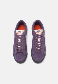 Ein Paar lila Wildleder-Nike-Sneaker mit schwarzen Sohlen, weißen Swoosh-Logos und orangen Innensohlen, von oben auf weißem Hintergrund betrachtet.