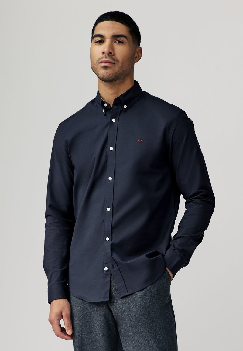 Camicia blu navy con bottoni, realizzata in cotone con colletto strutturato, bottoni bianchi e un piccolo emblema rosso sul petto.