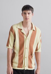 Kurze ärmelige, knöpfbare Strickjacke mit gelben und braunen Streifen, ausgestattet mit einem strukturierten Strickmuster, einem spitzen Kragen und zwei vorderen Taschen.