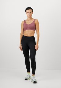 Kvinne som står iført en mauve trenings-BH, svarte høyhårede leggings med små perforeringer, og hvite joggesko med neongrønne sokker.