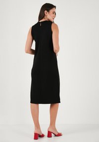 Black sleeveless dress with a straight cut, knee-length, featuring a small back keyhole and side slits, paired with red block-heeled sandals.