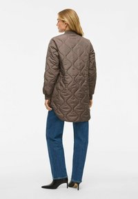 Vrouw in een lange, gewatteerde bruine jas, blauwe jeans en zwarte hakken enkellaarzen, staand met haar rug naar de camera op een witte achtergrond.