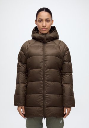 Une femme porte un long manteau matelassé marron à capuche, avec une fermeture éclair sur le devant et un logo, sur un fond clair uni.