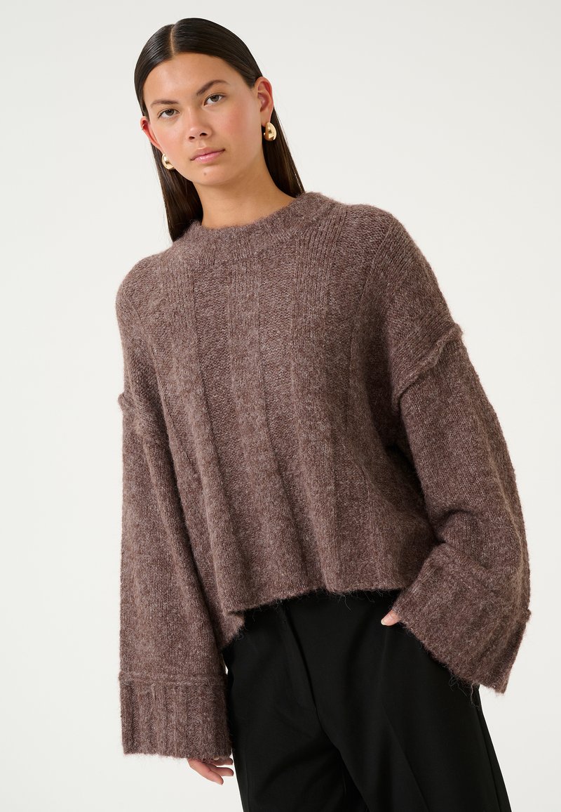 Brown knitted sweater with a relaxed fit, wide sleeves, ribbed cuffs, and a round neckline. Soft texture with visible stitch patterns.