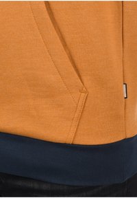Oranges Sweatshirtstoff mit strukturierter Oberfläche und navyfarbendem unteren Saum. Seitentaschendetails sichtbar, mit einem kleinen Etikettakzent.