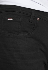 Vaqueros de denim negro con un bolsillo trasero que posee una pequeña etiqueta metálica con la inscripción "Petrolino" y detalles de remaches de cobre. Textura suave.