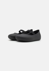 Scarpe nere piatte in gomma flessibile, con punta arrotondata e due cinturini elastici sulla parte superiore per una vestibilità sicura. Suola testurizzata per una buona aderenza.