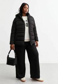 Svart puffjacka med huva, kviltad textur, parat med en vit sweatshirt, vida svarta byxor och krämfärgade sneakers, medan hon håller en svart handväska.