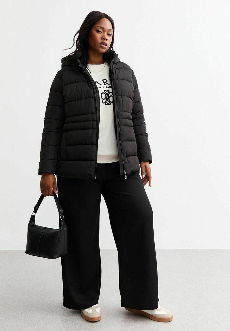 Svart puffjacka med huva, kviltad textur, parat med en vit sweatshirt, vida svarta byxor och krämfärgade sneakers, medan hon håller en svart handväska.