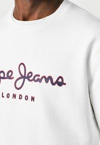 En person som bär en vit sweatshirt med "Pepe Jeans London" broderat i marinblått och rött på bröstet.