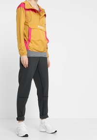 Giacca leggera gialla e rosa con tasca frontale, camicia grigia a strati, pantaloni neri a gamba tapered e scarpe da ginnastica bianche. Texture liscia.