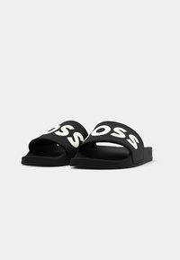 Paire de sandales noires à enfiler avec un texte blanc en gras sur les brides, sur un fond gris clair uni.