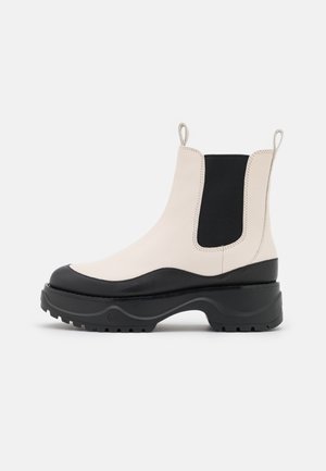 Bottines Chelsea en cuir crème et noir avec semelle épaisse à crampons, panneaux latéraux élastiques et languettes sur fond blanc, vue latérale.