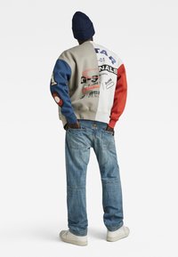 Kleurrijke sweatshirt met grijze, blauwe en rode mouwen; bevat logo's en tekst. Gepaarde met lichtblauwe denimjeans en crème sneakers.