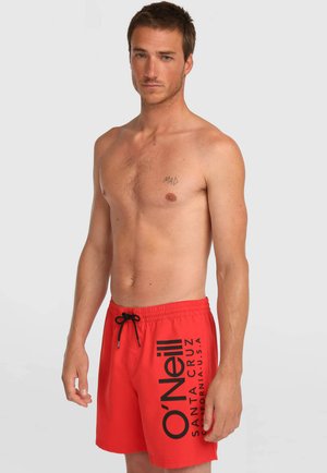 Homme torse nu aux cheveux bruns courts, portant un short de bain rouge O'Neill avec un cordon noir, debout devant un fond clair uni.