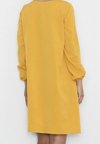 Gelbes Kleid mit langen Ballonärmeln, rundem Ausschnitt und lockerer Passform. Aus glattem Stoff gefertigt, fällt es über das Knie.