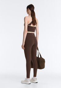 Femeie cu părul lung, purtând un sutien sport maro și colanți, adidași albi, ținând o geantă de sală maro închis, cu fața spre spate pe un fundal simplu.