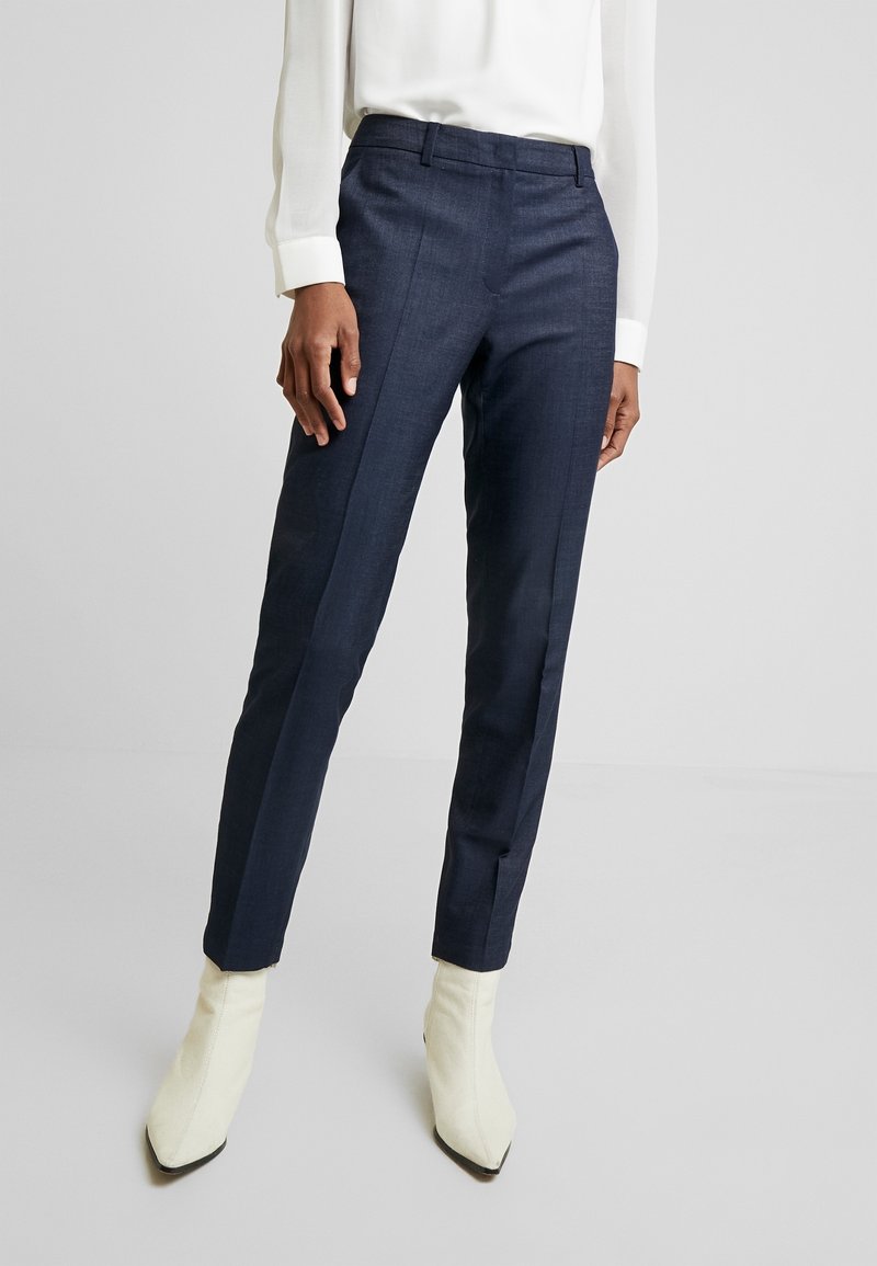 Pantalon bleu marine sur mesure en tissu texturé avec une coupe slim, associé à des bottines crème et un chemisier blanc.