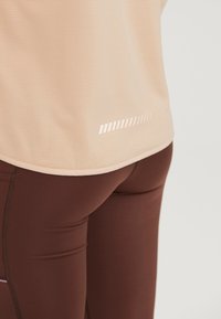 Lys beige athletic top med en tekstureret overflade og refleksbånd. Parret med brune tætsiddende leggings, der fremviser et glat, strækbart stof.