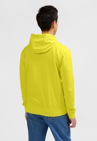 Jugend-Hoodie in leuchtendem Gelb mit einfarbigem Design, langen Ärmeln und Kapuze mit Kordelzug. Kombiniert mit blauen Denim-Jeans.