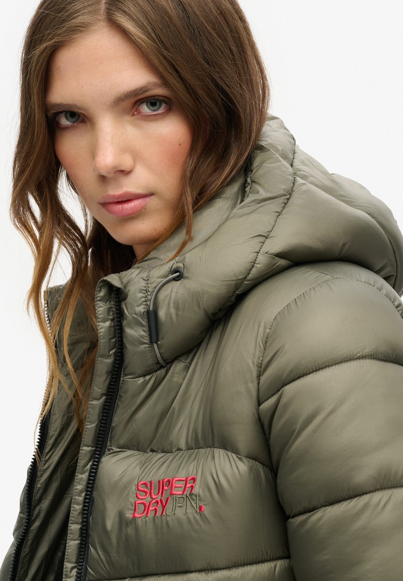 Olijfgroene pufferjack met hoge kraag, ritssluiting en verstelbare trekkoordkap. Voorzien van een rood geborduurd "SUPERDRY" logo.