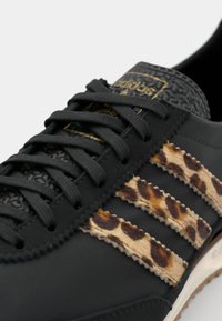 Black athletic shoe featuring leopard print accents on three stripes, textured upper, and black laces. Gold branding present on the tongue.