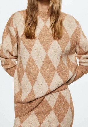Femme portant un ensemble pull et jupe beige et brun clair à motif losanges, debout les mains derrière le dos, cheveux longs et raides visibles.