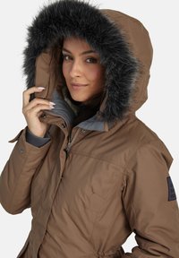 Bruine geïsoleerde parka met een zwarte nep bonten capuchon, ritssluiting aan de voorkant en drukknoopsluiting. Bevat zijzakken en elastische manchetten.