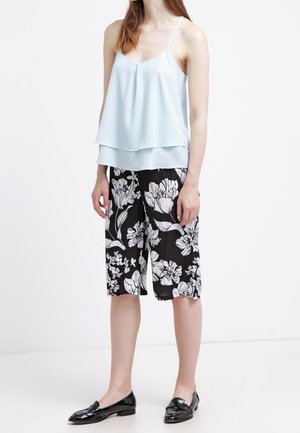 Vrouw die een lichtblauw mouwloos topje draagt, zwarte knielange shorts met witte bloemenprint en zwarte loafers, staand tegen een witte achtergrond.