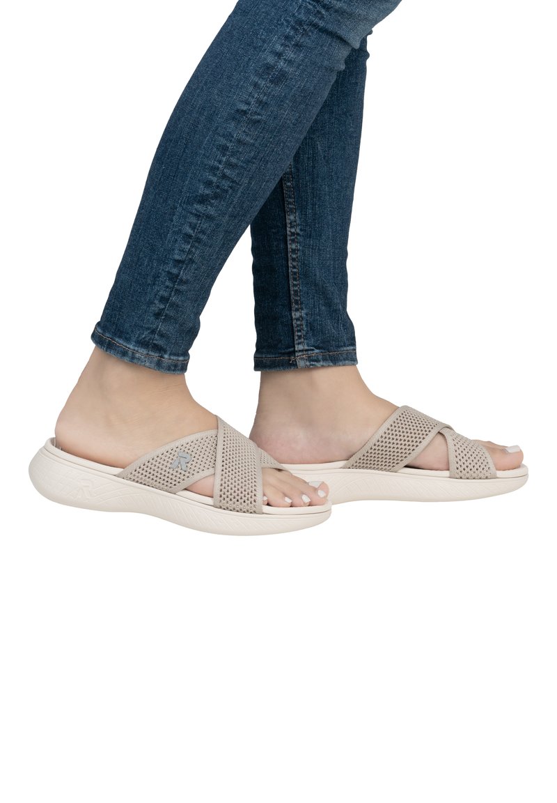 Light beige slip-on sandals feature crisscrossed perforated straps and a textured white sole, designed for comfort. Worn with dark blue jeans.