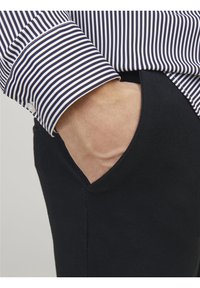 Personne portant un pantalon noir avec la main dans la poche et le revers d'une chemise à manches longues rayée noir et blanc visible.