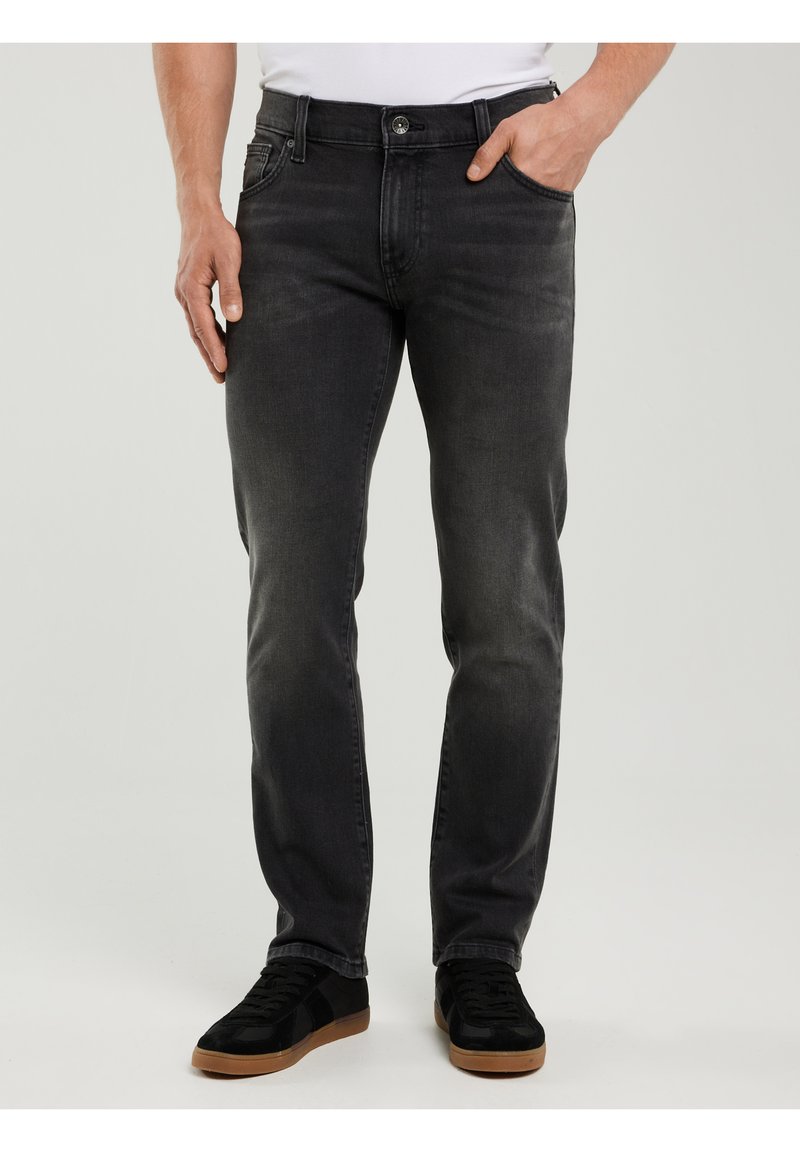 Mężczyzna w wyblakłych czarnych dżinsach, w białej koszuli wpuszczonej do spodni, z jedną ręką w kieszeni oraz czarnych sneakersach z gumowymi podeszwami.