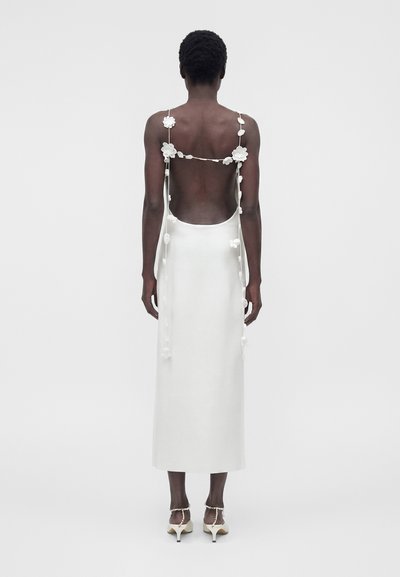 Γυναίκα με κοντά μαλλιά που φοράει λευκό φόρεμα χωρίς πλάτη με λουλουδένιους ιμάντες και λευκά ψηλοτάκουνα παπούτσια, στέκεται μπροστά σε απλό φόντο.