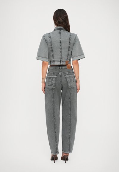 Kvinne med langt mørkt hår iført en gråvasket cropped jakke og høytlivde jeans med blokkbokstavene "B" og "P" på baklommene, stående.