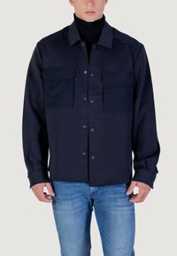 Man in een donkerblauwe button-up jas met twee borstzakken over een zwarte coltrui en lichtblauwe jeans.