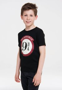 Black cotton t-shirt featuring a circular white graphic with red accents and the text "PLATFORM 9¾." Paired with dark pants.