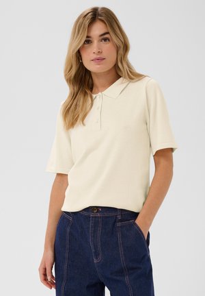 Krém színű galléros póló, klasszikus nyakkal, rövid ujjakkal és négy gombbal. Az anyag puhanak tűnik, enyhe textúrával.