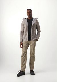 Casaco leve cinzento com fecho de correr sobre uma camisa preta, combinado com calças cor de castanho claro com bolsos. Completa o conjunto com sapatos pretos com detalhes vermelhos.