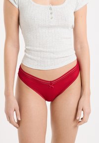 Mutandine rosse a coste con rifiniture di pizzo e dettaglio a fiocco. Tessuto morbido e fascia elastica in vita per il comfort. Abbinate a una camicia grigia chiaro con bottoni.