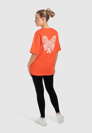 T-shirt en coton orange à manches courtes avec un grand graphique de papillon blanc dans le dos. Associé à des leggings noirs et des baskets blanches.