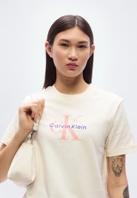 Κρεμ βαμβακερό T-shirt με μεγάλο ροζ και μπλε λογότυπο Calvin Klein. Το μοντέλο κρατάει μια μικρή λευκή τσάντα χειρός με καμπυλωτό σχήμα.