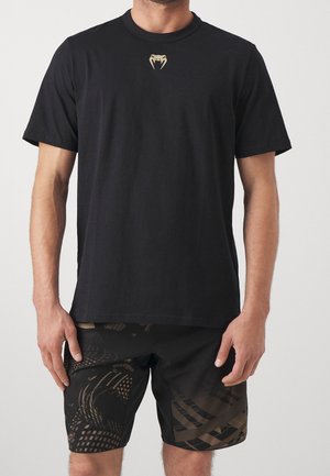 Man draagt een effen zwarte korte mouw T-shirt met een klein beige slangtandlogo in het midden van de borst en zwarte short met patroon.