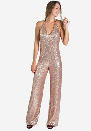 Combinaison en sequin rose gold avec un col halter, des jambes larges et une texture scintillante, mettant en avant un décolleté en V profond et des bretelles minimalistes.
