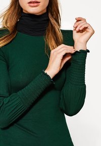 Musta tummanvihreä pitkähihaine paita, jossa on ribbikuvio. Fitted-mallinen, rypytetty yksityiskohta hihansuissa, ja korkea musta poolopaita alla.