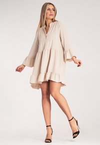 Beżowa sukienka w kształcie litery A z kołnierzykiem, trzema ćwierć rękawami w kształcie dzwonu i falbaniastym dołem. Wykonana z lekkiego materiału, z teksturowaną panelą.