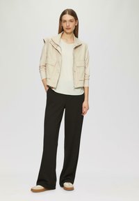 Beige mouwloze jas met voorkeurszakken, gelaagd over een licht topje, gecombineerd met zwarte wijde broek en beige sneakers. Glad textuur overal.