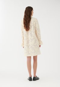 Vestido de crema claro con patrón de encaje floral, mangas largas con puños acampanados y un corte holgado. Llevado con mocasines negros. Fondo liso.