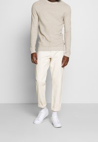 Man wearing beige ribbed sweater, off-white rolled-up pants, white socks, and white sneakers standing against a plain light background.